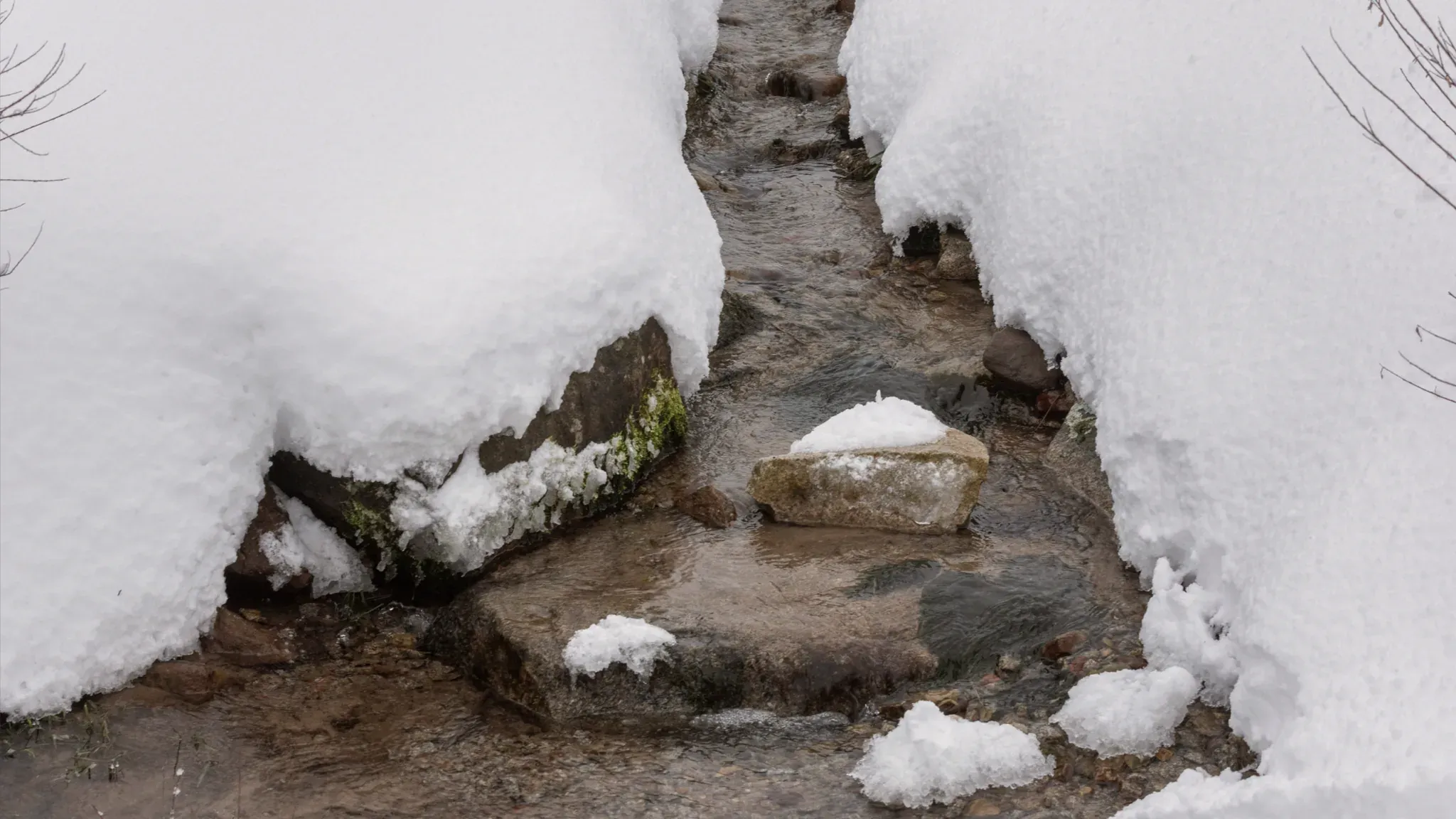 Ein schmaler Bach fließt zwischen schneebedeckten Ufern über Steine.