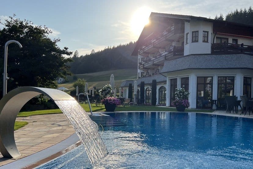 Außenpool des Engel Obertal bei Sonnenuntergang mit Schwall-Wasserfall, dahinter das Hotelgebäude und die Schwarzwaldhänge.