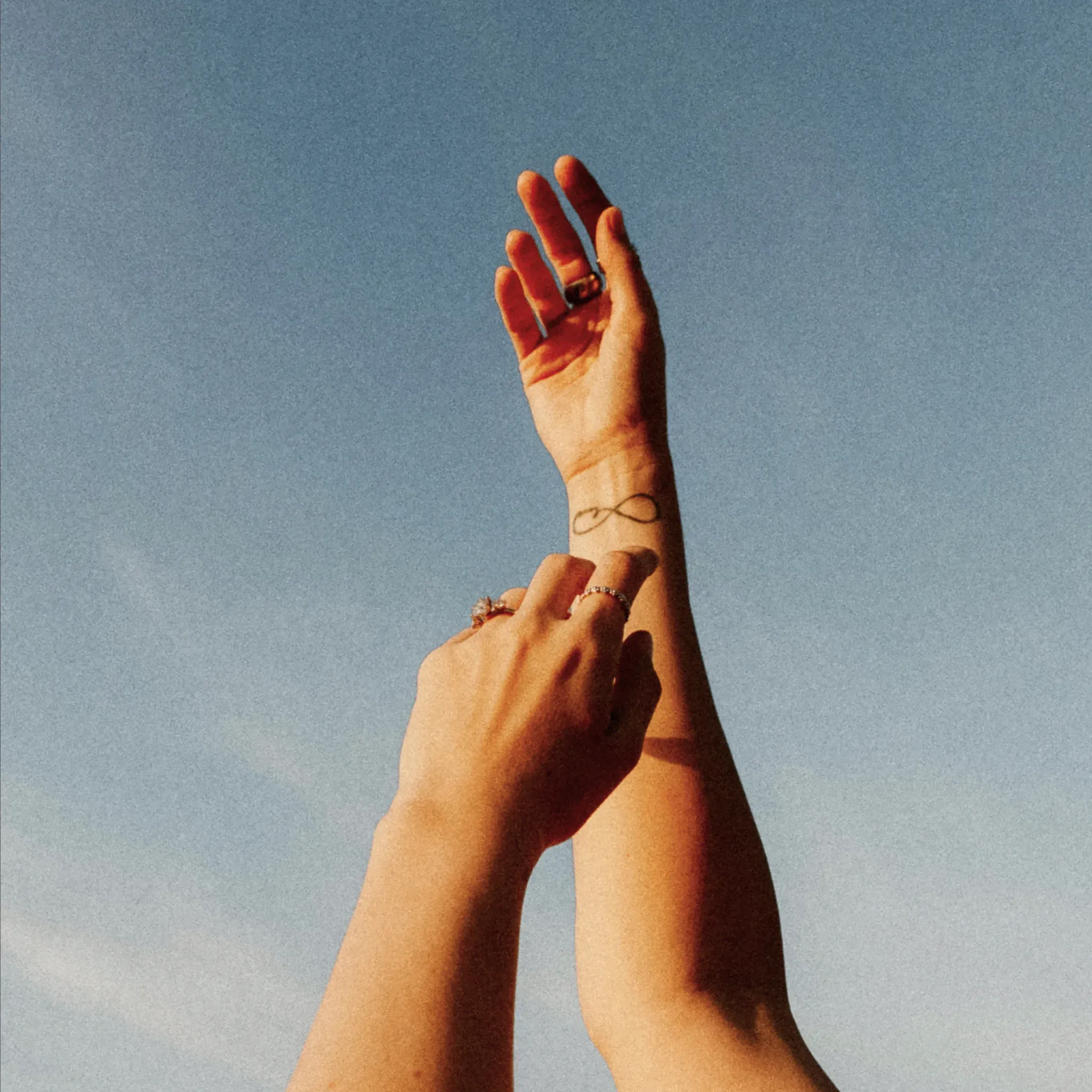 Zwei Hände berühren sich vor blauem Himmel, am Handgelenk ein Unendlichkeitstattoo.