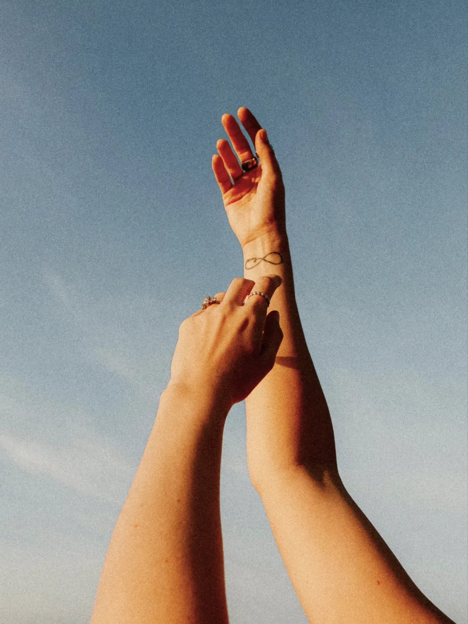 Zwei Hände berühren sich vor blauem Himmel, am Handgelenk ein Unendlichkeitstattoo.
