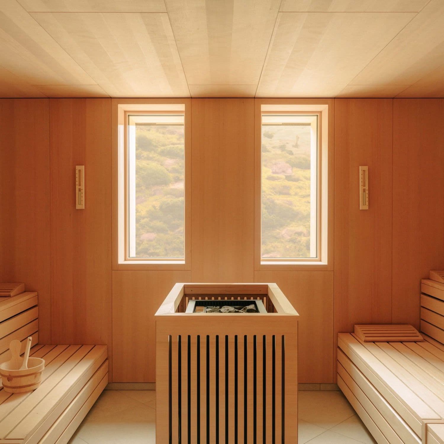 Holzverkleidetes Sauna-Interieur mit zwei hohen Fenstern zu grünen Hügeln und einem kleinen Holzkübel auf der Bank