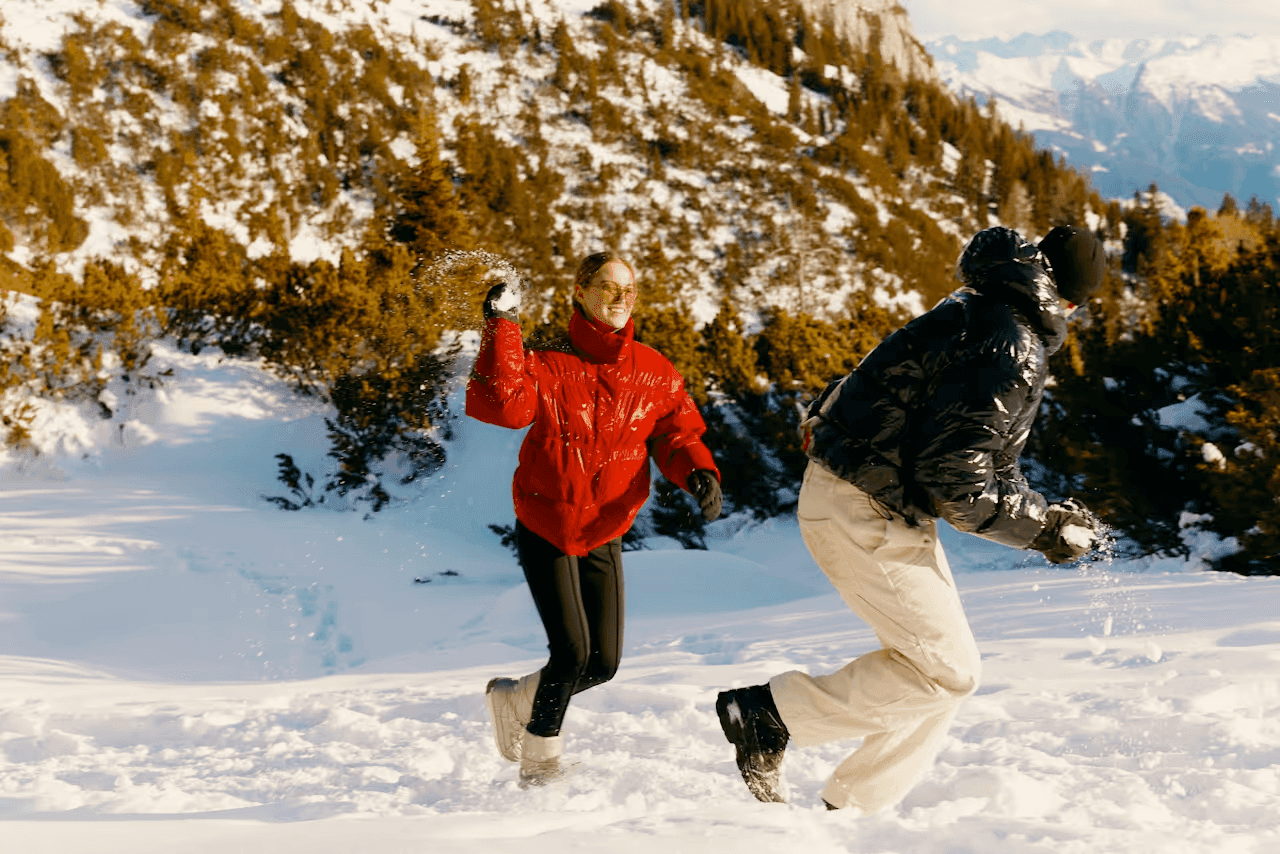 Paar in roten und schwarzen Daunenjacken bei einer Schneeballschlacht auf einem sonnigen Alpenhang