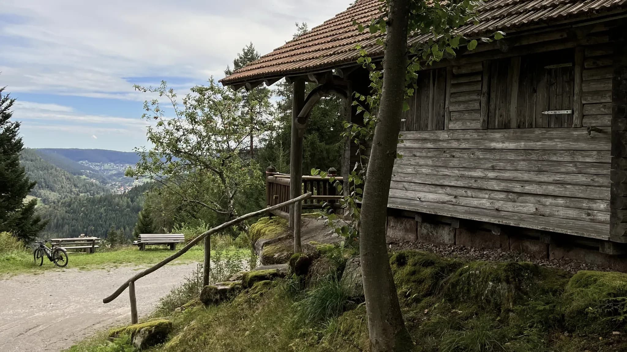 Verwitterte Holzhütte mit Vordach am Wanderweg, dahinter der Blick ins bewaldete Tal.