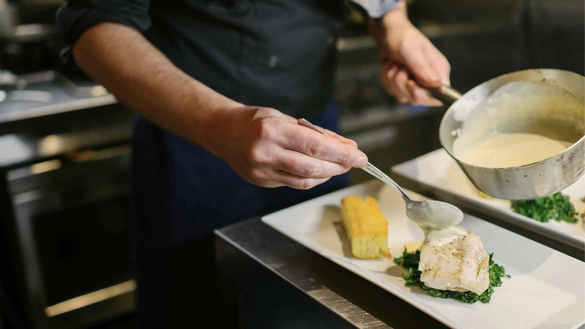 Koch richtet Fisch und Beilagen auf einer länglichen Platte an und gibt Sauce aus einer Kasserolle dazu.