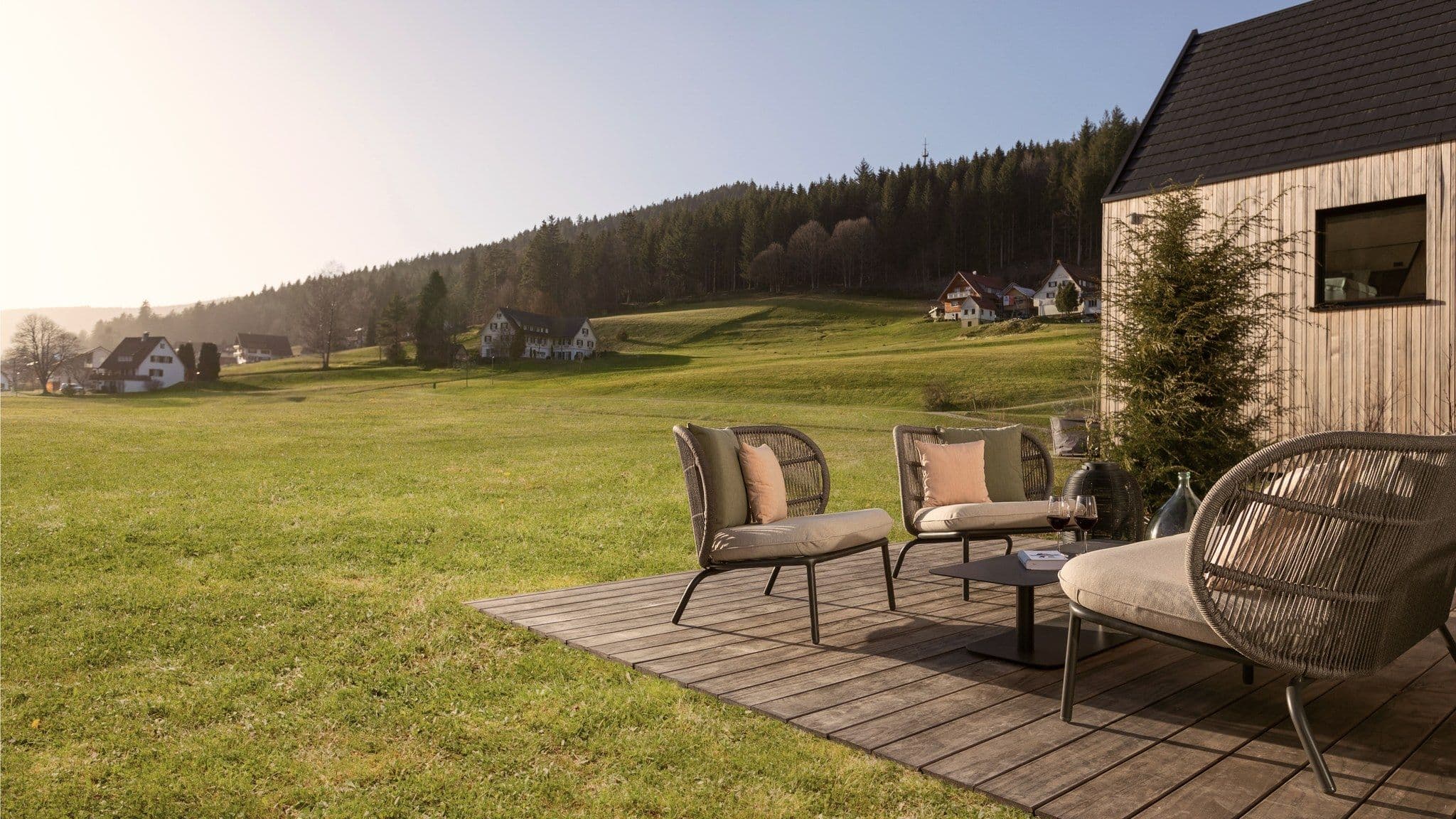 Holzterrasse eines Chalets mit Loungesesseln, davor Wiese und Schwarzwaldhang im Morgenlicht