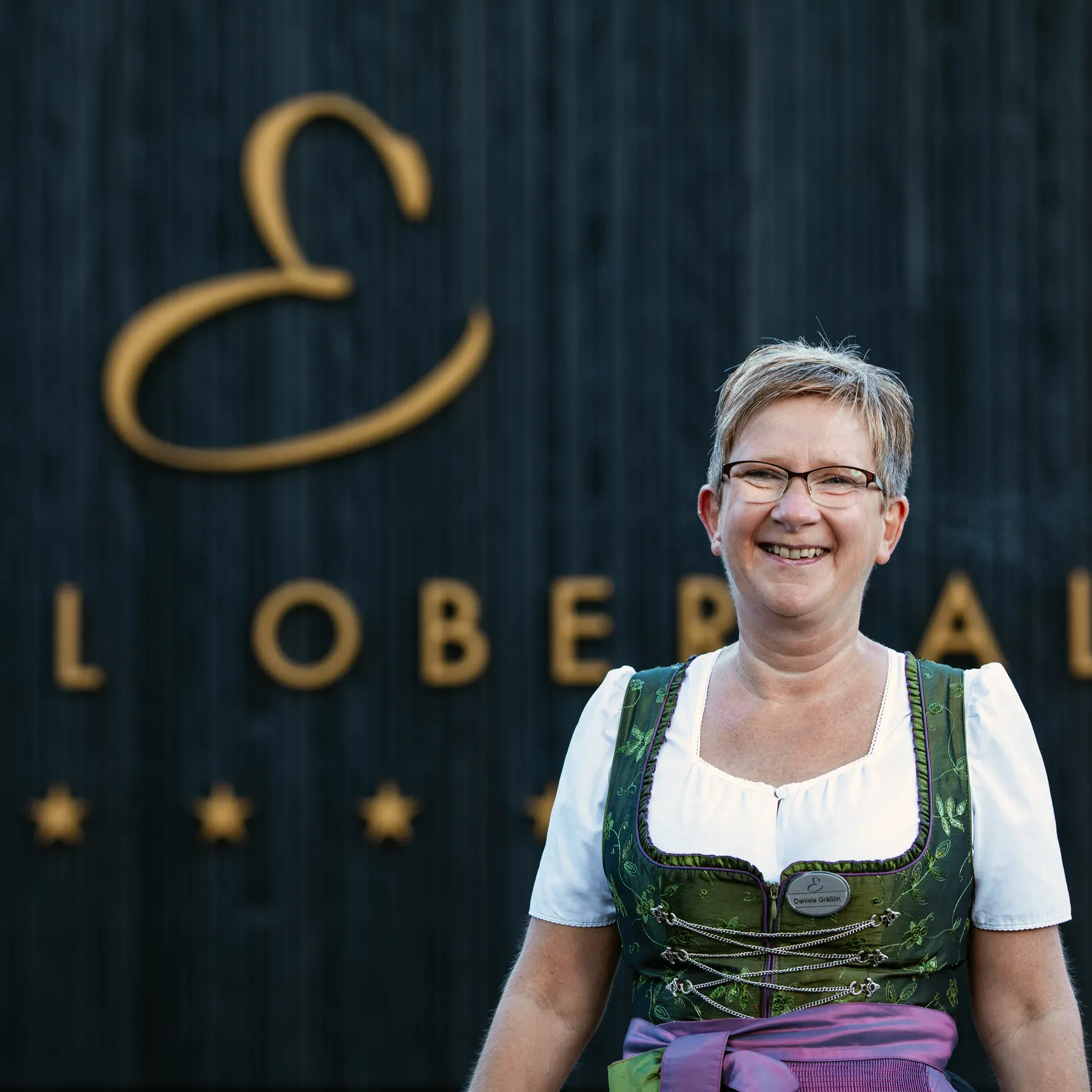 Porträt von Daniela Gräßlin, Restaurantleiterin im Engel Obertal, im Dirndl vor dunkler Holzwand mit goldenem Schriftzug.