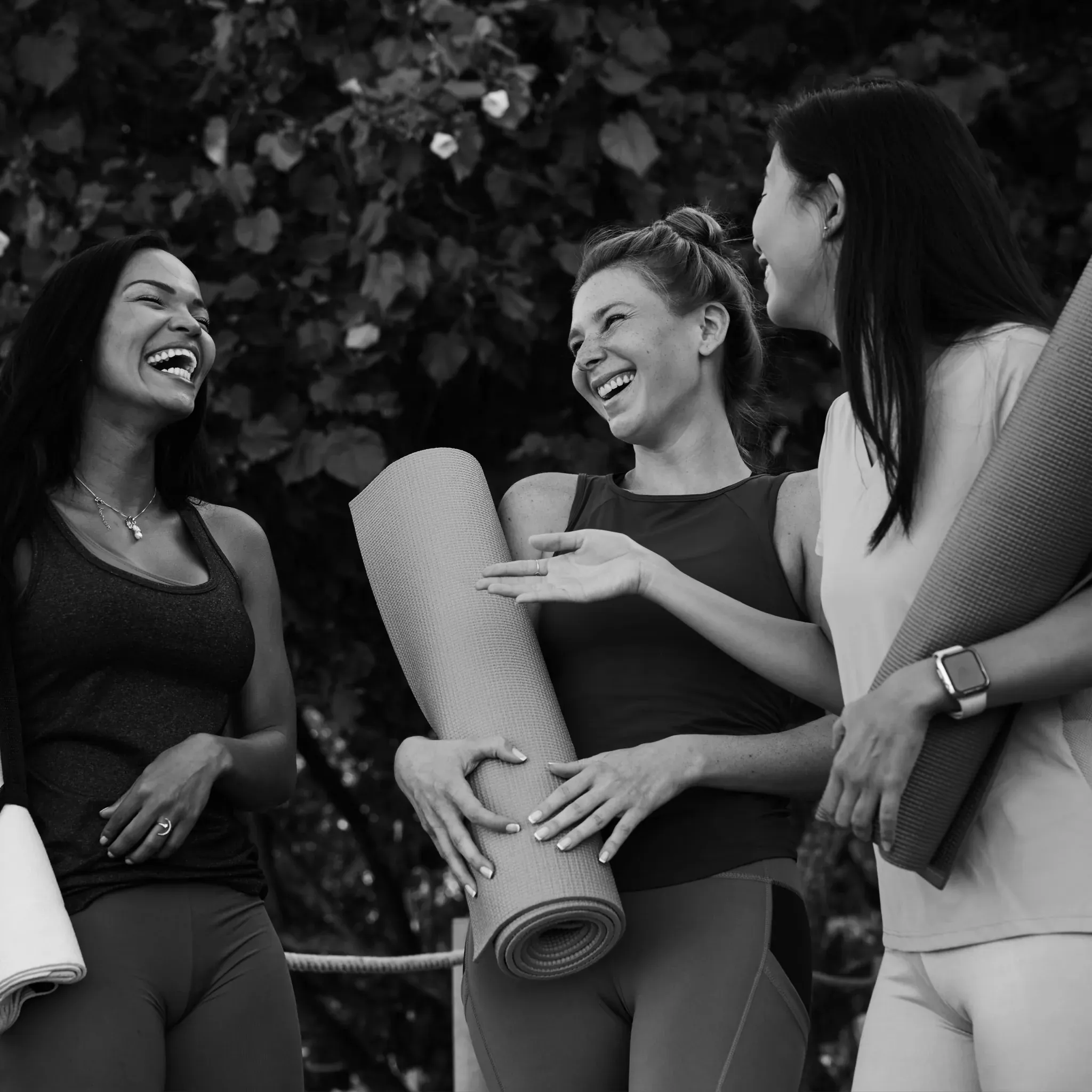 Drei lachende Frauen mit Yogamatten unter dem Arm im Gespräch, Schwarzweiß.