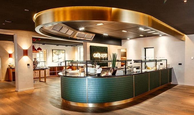 Geschwungene Buffettheke mit petrolfarbenen Fliesen und Messingrand unter goldfarbener Deckenverkleidung, im Hintergrund ein „Grüß Gott"-Schriftzug.
