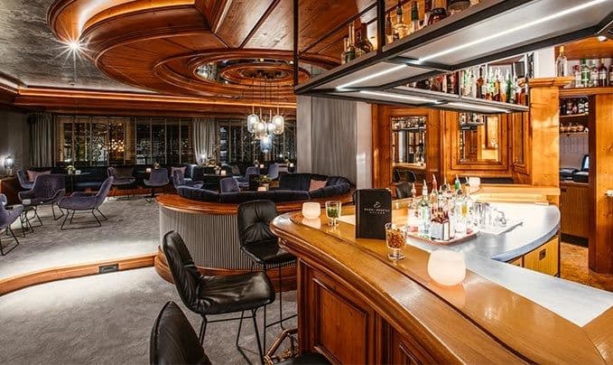 Hotelbar mit geschwungener Holzdecke, schwarzen Lederstühlen und bestücktem Flaschenregal.