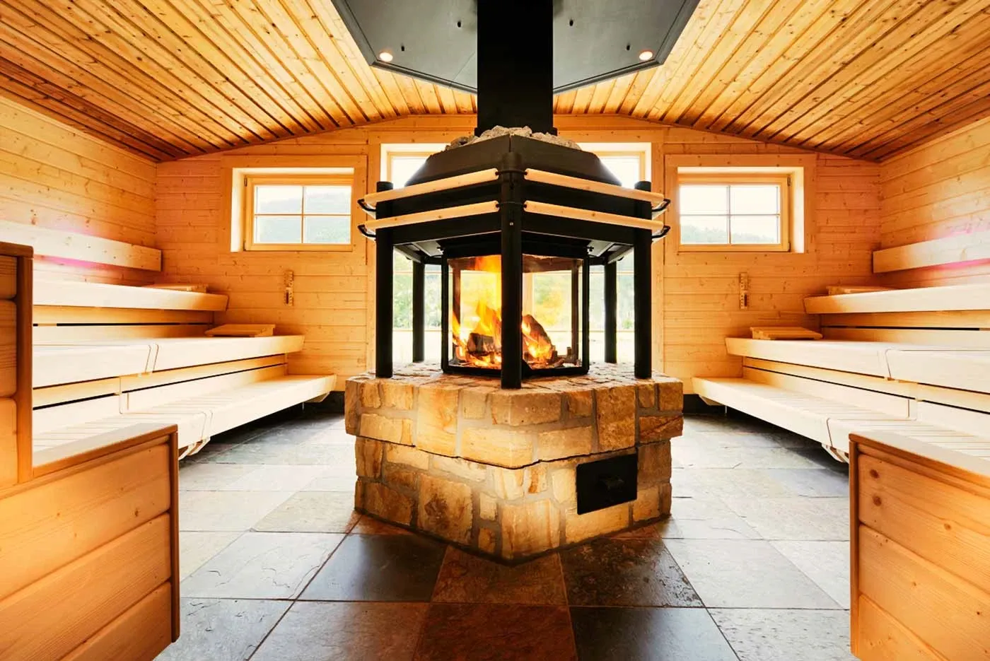 Holzvertäfelte Sauna mit umlaufenden Bänken und einem gemauerten, zentralen Kaminofen mit sichtbarem Feuer.