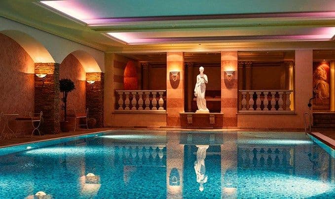 Beheiztes Innenbecken im Engel Obertal mit türkis schimmerndem Wasser, mediterranen Rundbögen und einer klassischen Statue im warmen Abendlicht.