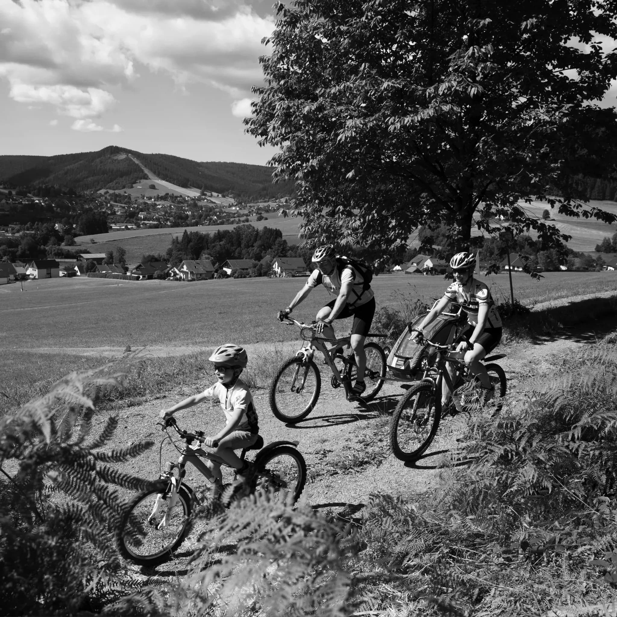 Schwarzweiß-Aufnahme einer Familie, die mit Mountainbikes einen Wiesenweg über dem Dorf hinauffährt.