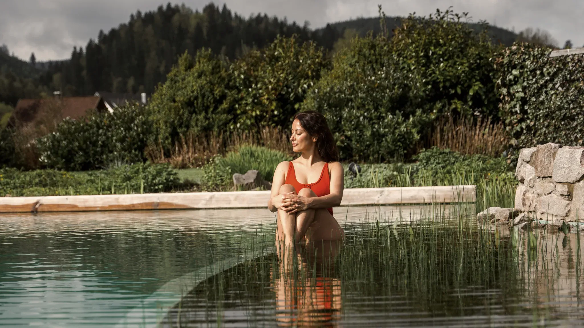 Frau in rotem Bikini sitzt im Wasser am Rand eines Naturbadeteichs vor bewaldeten Hügeln.