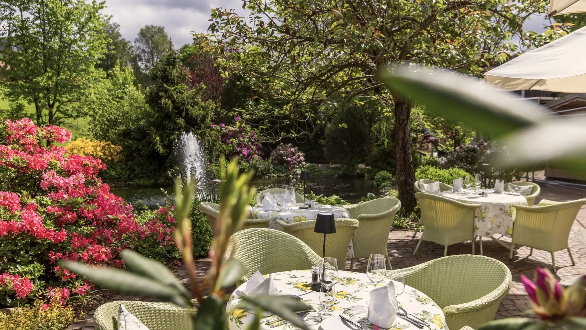 Gedeckte Tische mit hellgrünen Korbsesseln auf der Gartenterrasse, dahinter ein Springbrunnen und blühende Sträucher.