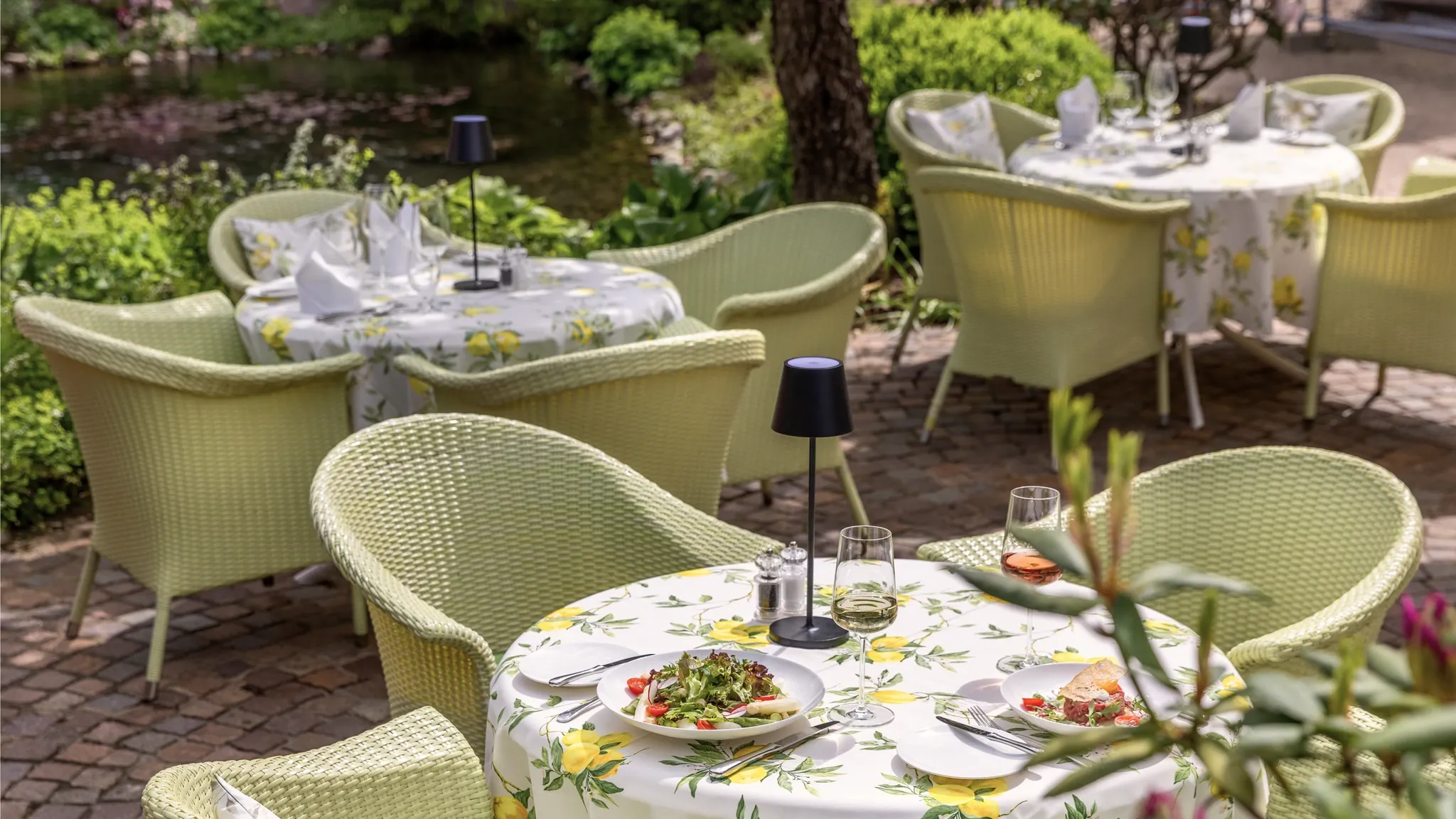 Gedeckter Tisch mit hellgrünen Korbsesseln auf der Gartenterrasse unter Bäumen.