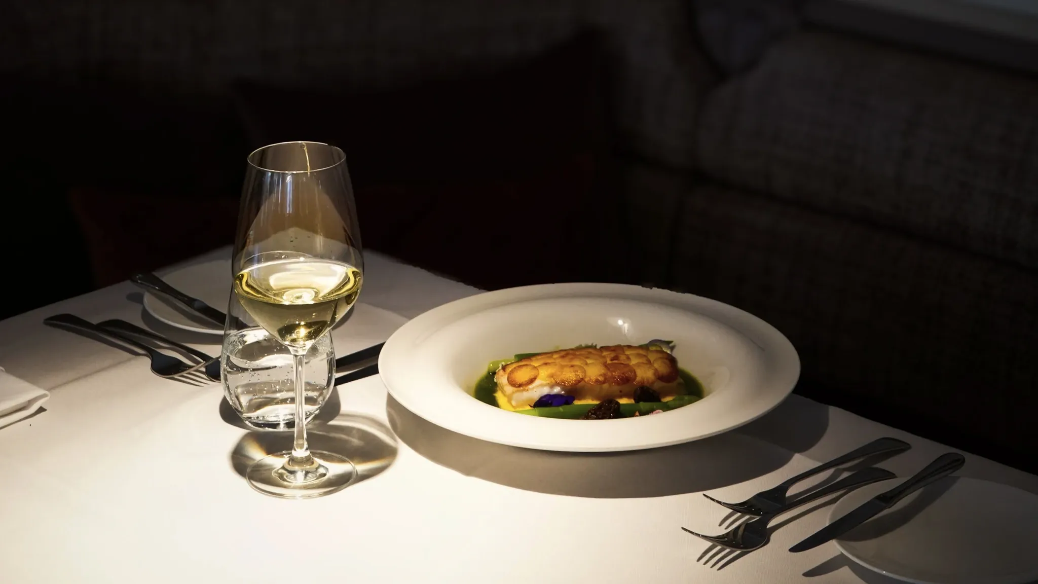 Weißer Teller mit Fischgericht und ein Glas Weißwein auf einem dunkel eingedeckten Restauranttisch im warmen Abendlicht.
