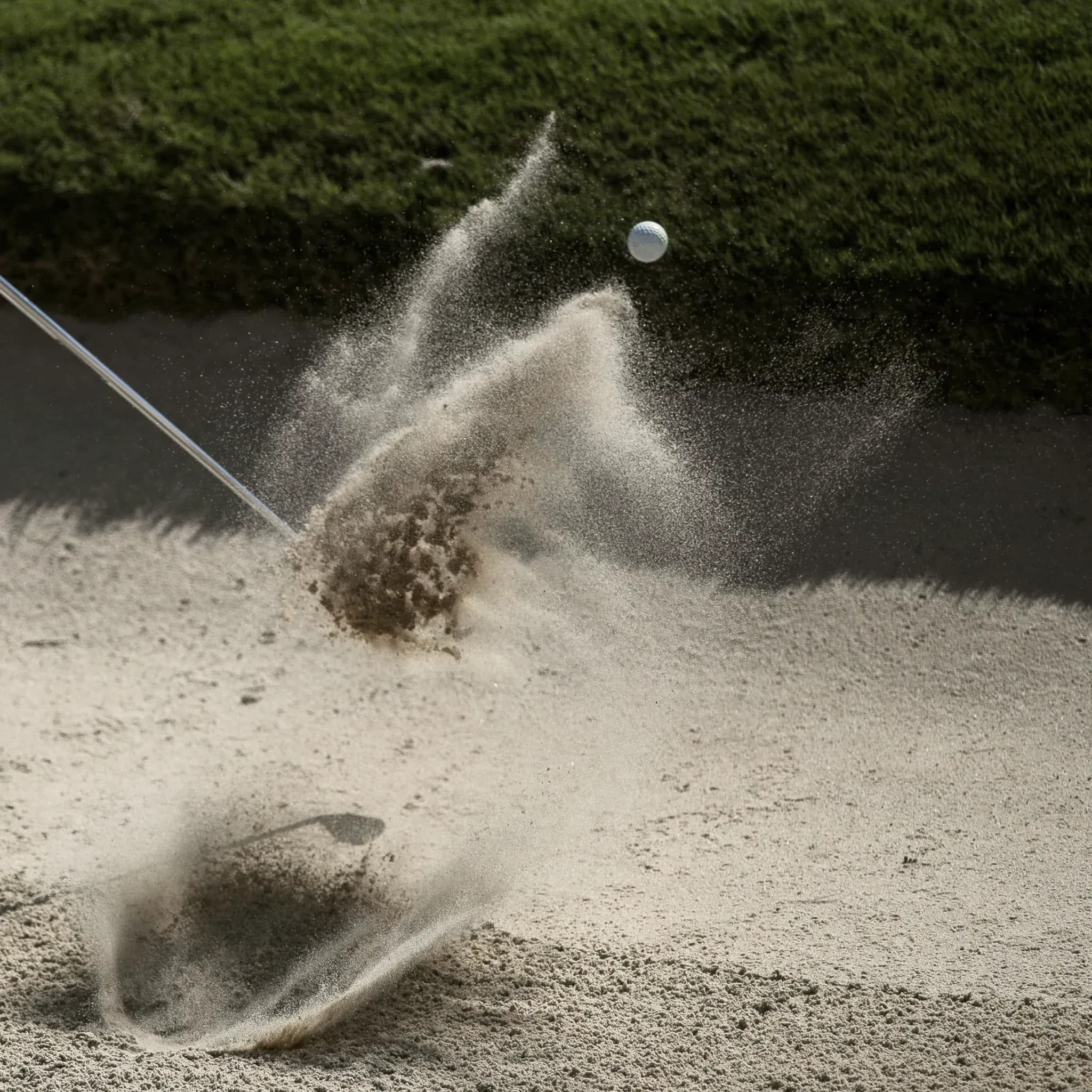 Golfball fliegt aus einem Sandbunker, aufgewirbelter Sand im Gegenlicht.