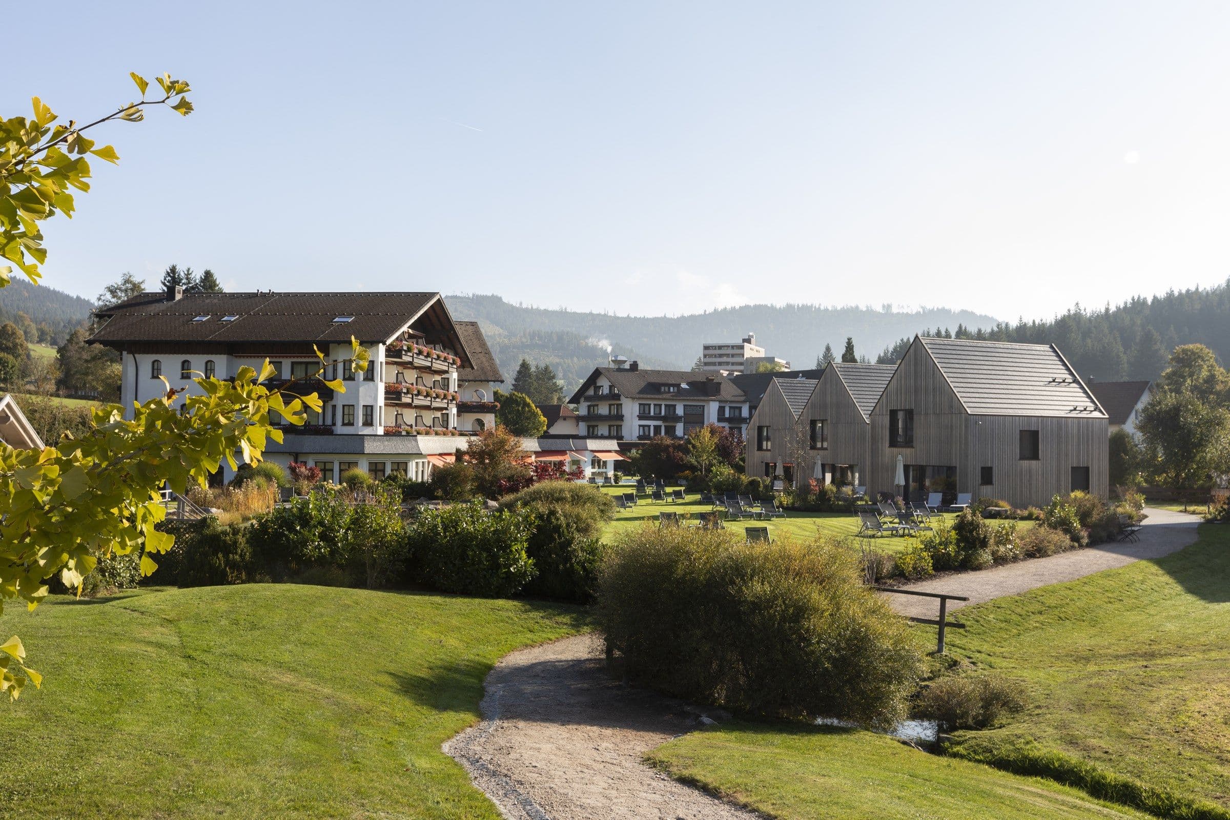 Blick über die Gartenanlage des Engel Obertal mit Bachlauf und Wiese auf das Haupthaus mit Blumenbalkonen und die modernen Chalets, Herbststimmung.
