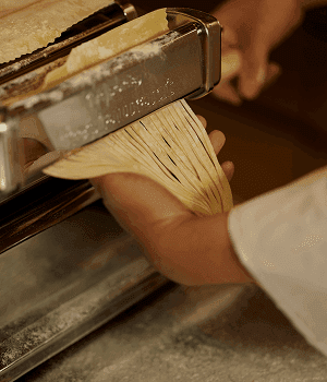 Hände eines Kochs führen frische Pastabahnen durch eine handbetriebene Nudelmaschine