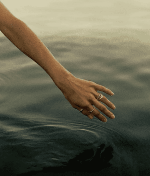 Eine Hand taucht sanft in die ruhige, dunkle Wasseroberfläche eines Sees ein.