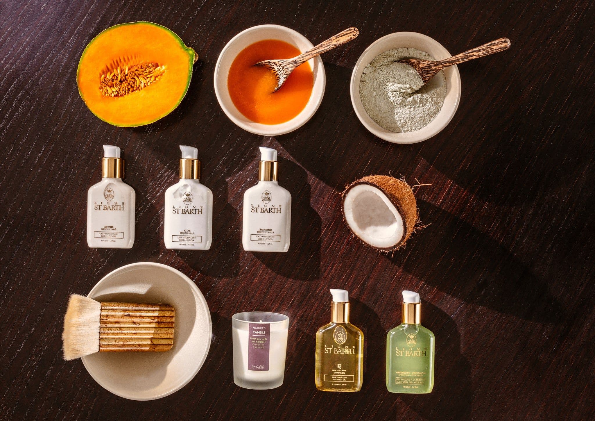 Flatlay auf dunklem Holz: Body-Lotionen, Bräunungsöl, Aloe-Gel und Duftkerze von Ligne St Barth, dazu halbierte Melone, Kokosnuss, Honig, Tonerde und ein Kosmetikpinsel.