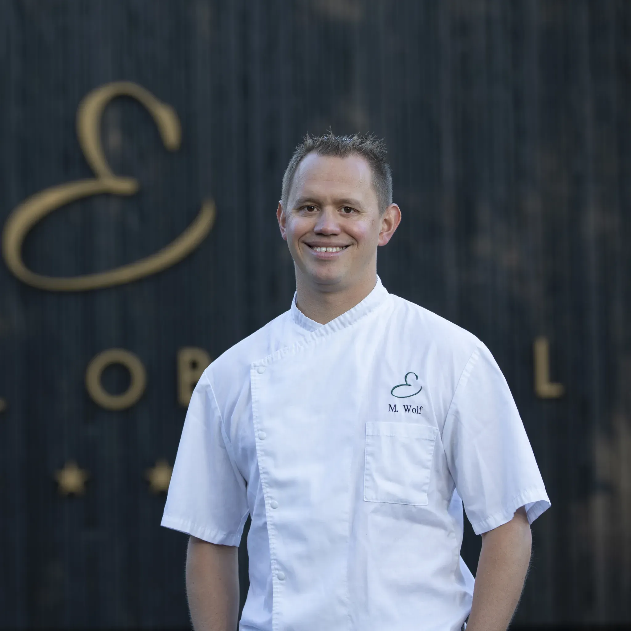 Porträt von Markus Wolf, Küchenchef im Engel Obertal, in weißer Kochjacke vor dunkler Holzwand mit goldenem Schriftzug.