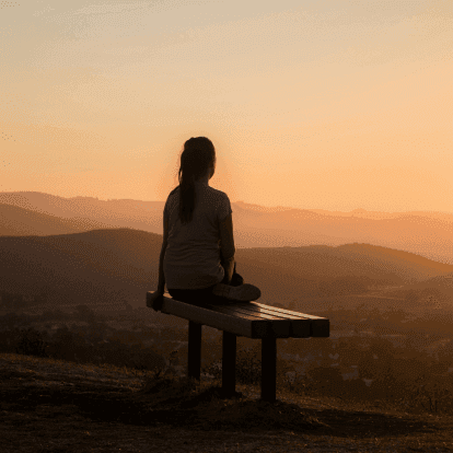 Silhouette einer Person, die auf einer Holzbank sitzt und den Sonnenuntergang über gestaffelten Bergketten betrachtet.