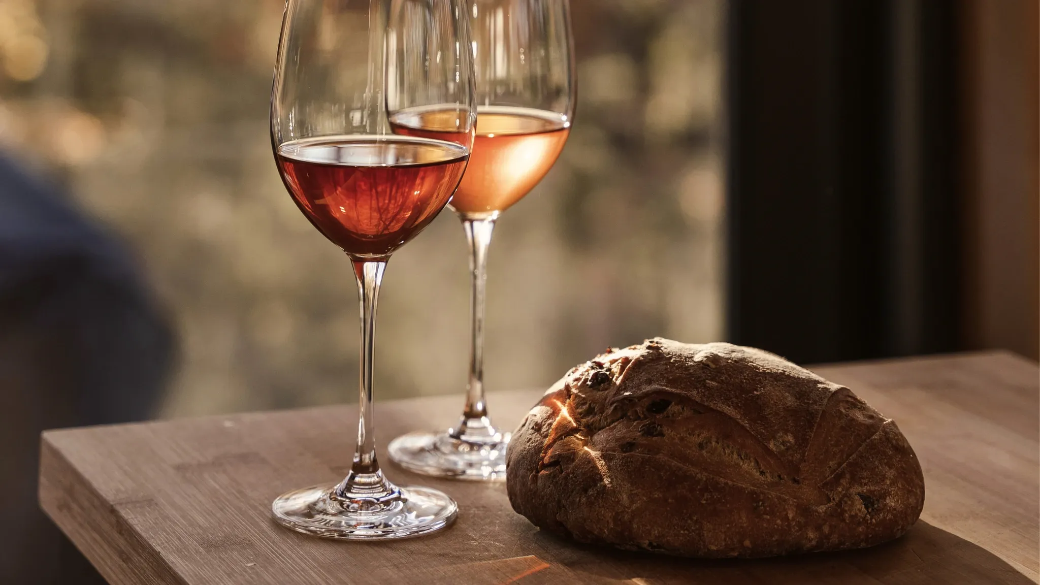 Zwei Gläser Roséwein und ein rundes Brot auf einer Holzplatte vor einer Natursteinwand.