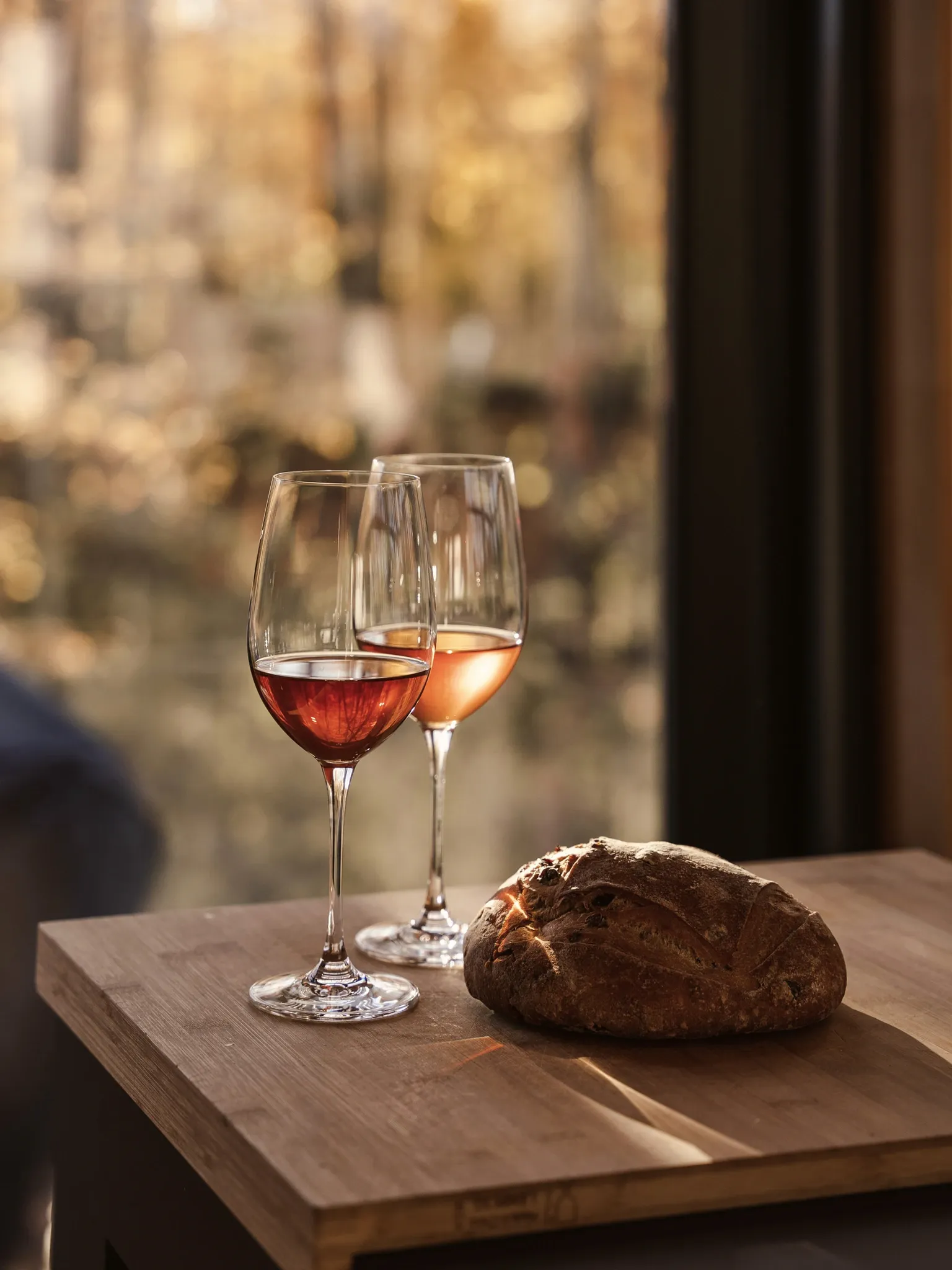 Zwei Gläser Roséwein und ein rundes Brot auf einer Holzplatte vor einer Natursteinwand.