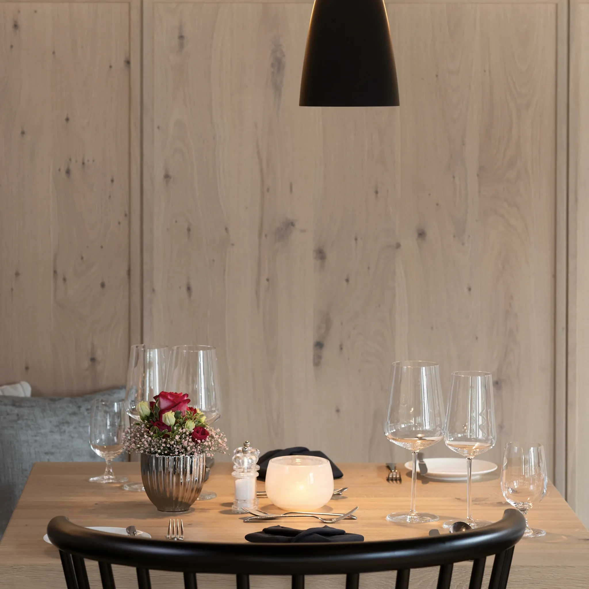 Runder Tisch mit Weingläsern, Blumen und Teelicht an einer hellen Holzwand unter einer schwarzen Pendelleuchte.
