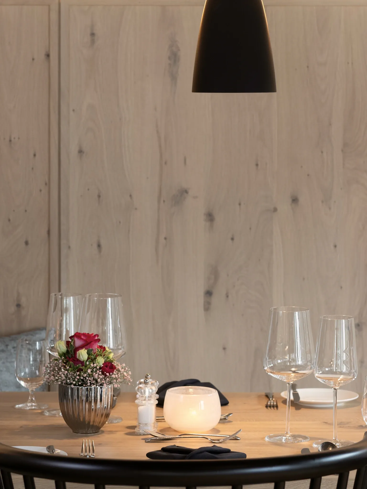Runder Tisch mit Weingläsern, Blumen und Teelicht an einer hellen Holzwand unter einer schwarzen Pendelleuchte.