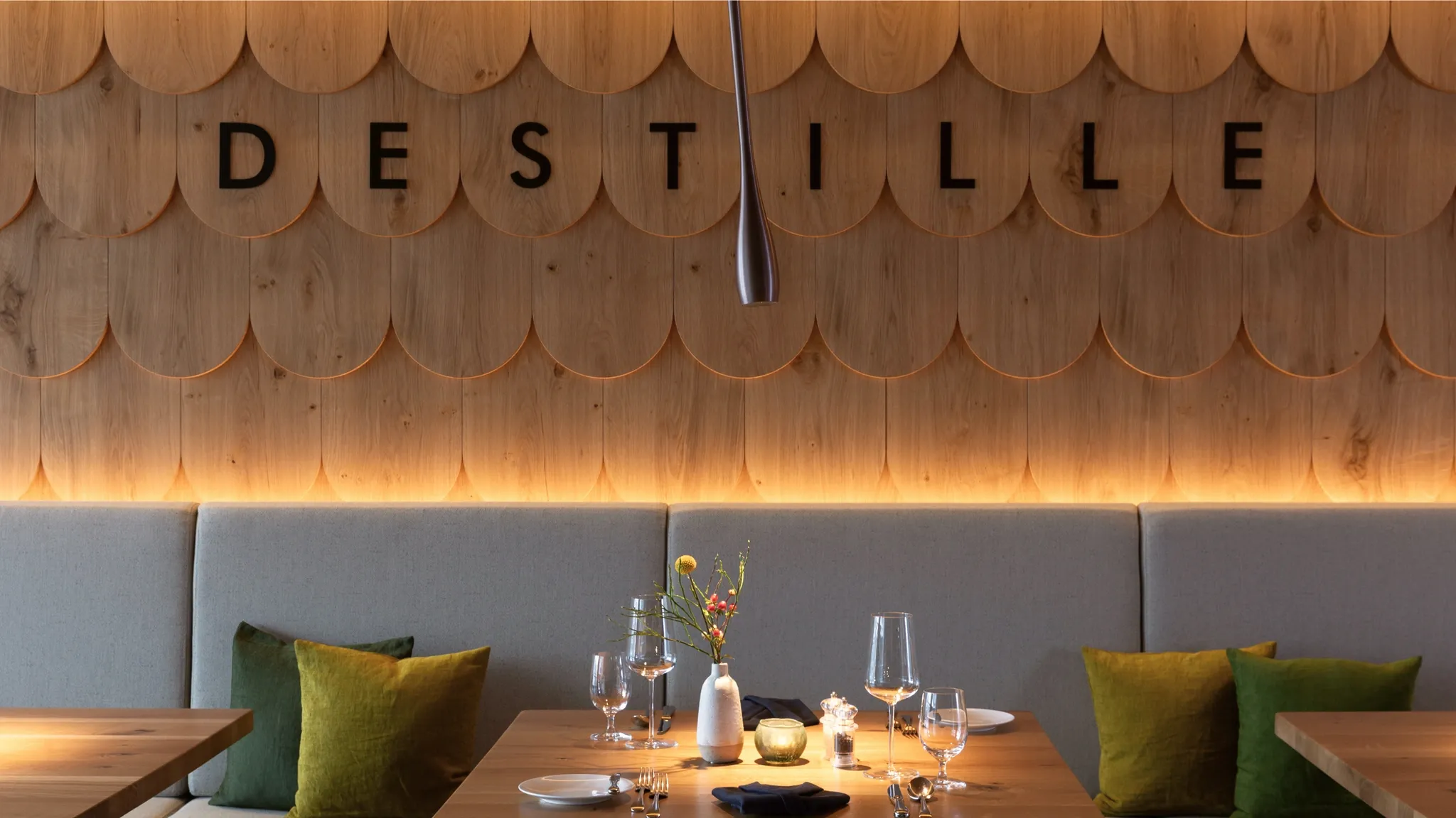 Holzschindelwand mit dem Schriftzug Destille, darunter eine gepolsterte Sitzbank und ein eingedeckter Tisch.