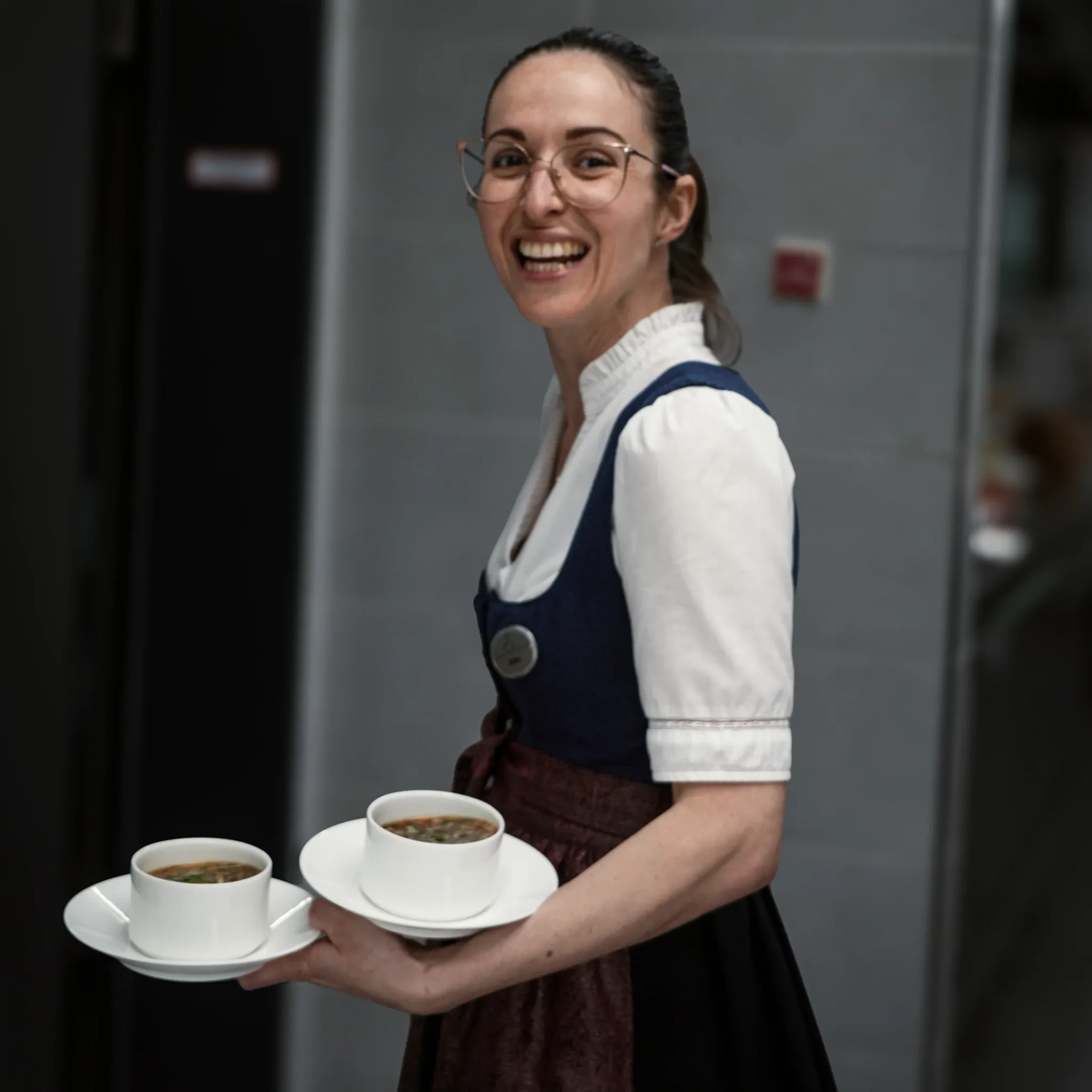 Lächelnde Servicemitarbeiterin im Dirndl trägt zwei Tassen auf Untertellern.