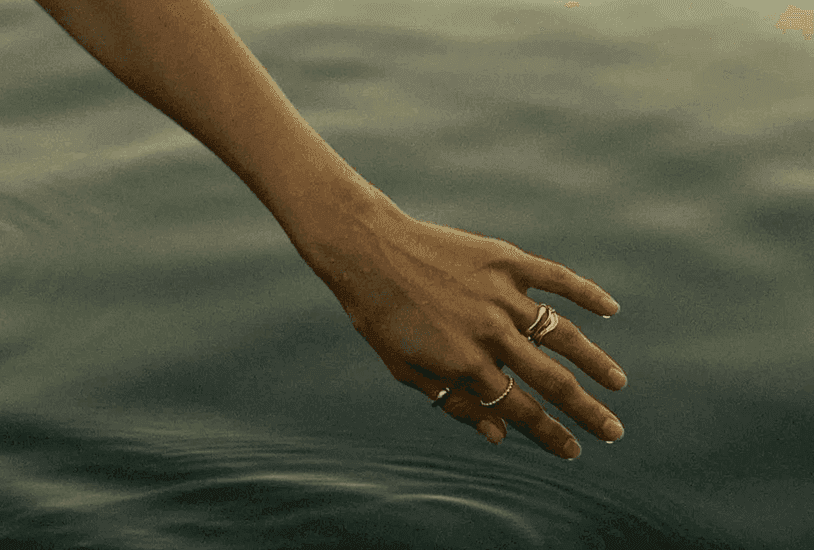 Hand einer Frau mit mehreren Silberringen berührt die Oberfläche von stillem, grünem Wasser