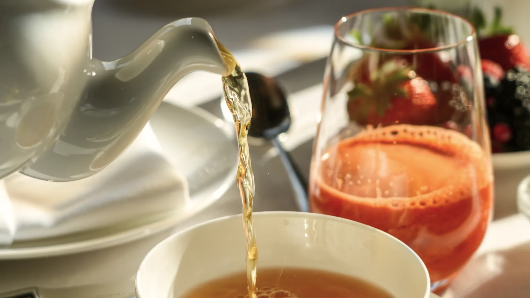 Tee wird aus einer Kanne in eine weiße Tasse gegossen, daneben ein Aperitif mit Erdbeeren.
