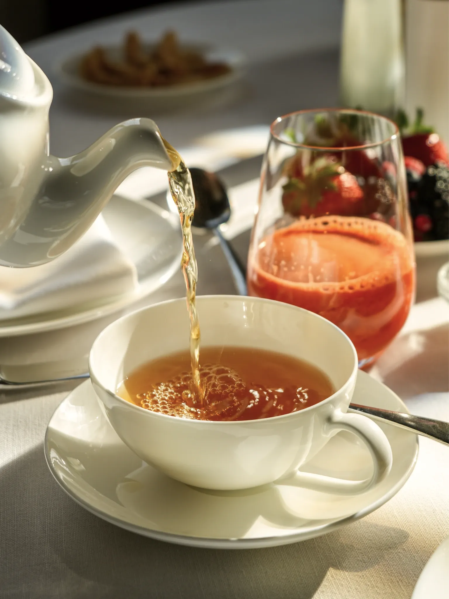 Tee wird aus einer Kanne in eine weiße Tasse gegossen, daneben ein Aperitif mit Erdbeeren.