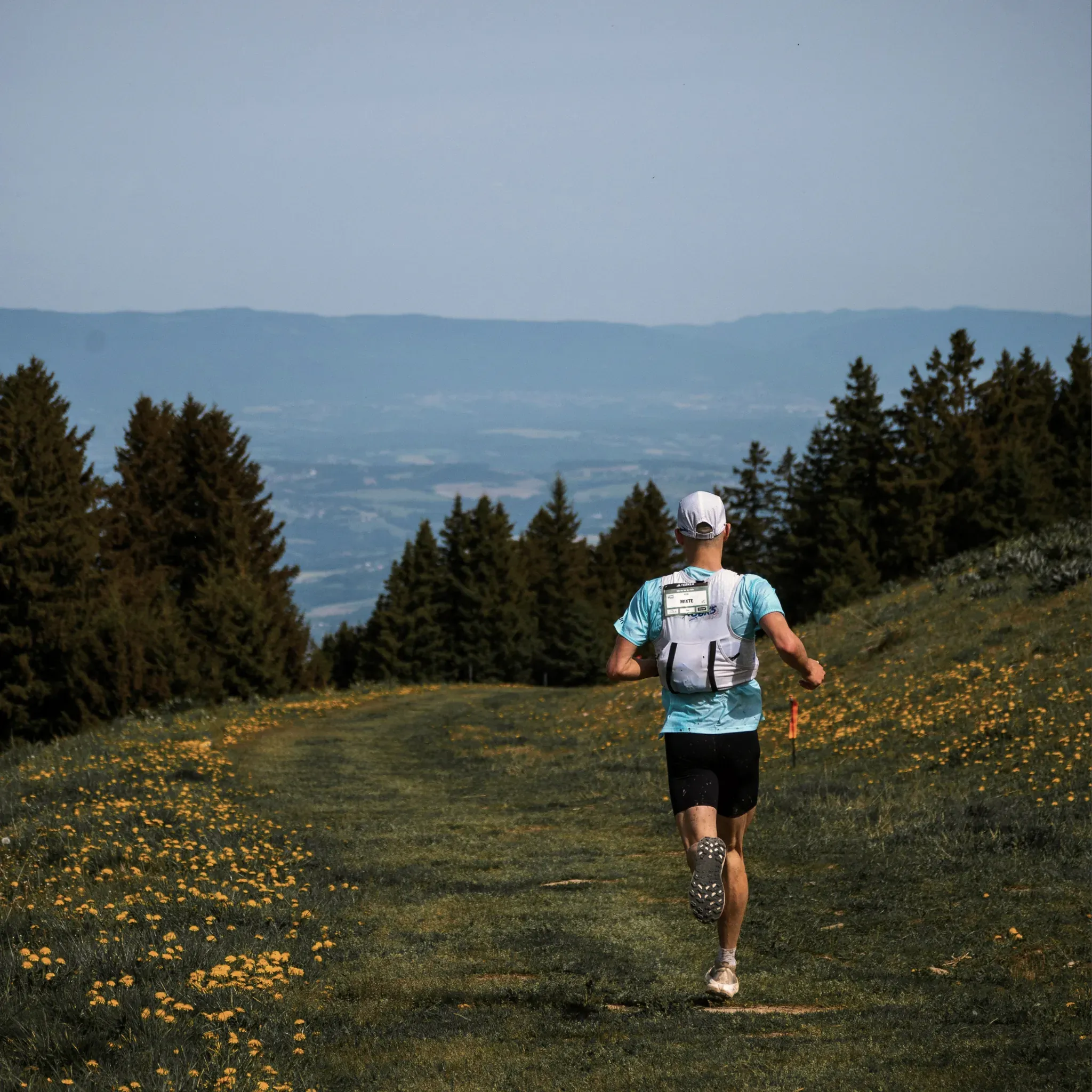 Läufer mit Trinkweste joggt über eine blühende Bergwiese am Waldrand.
