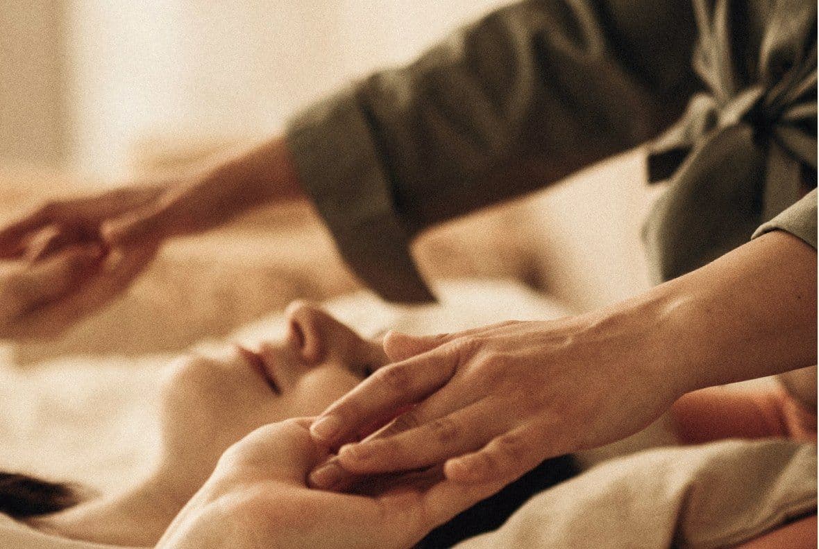 Kosmetische Behandlung: Hände einer Therapeutin an Kopf und Hand einer entspannt liegenden Person