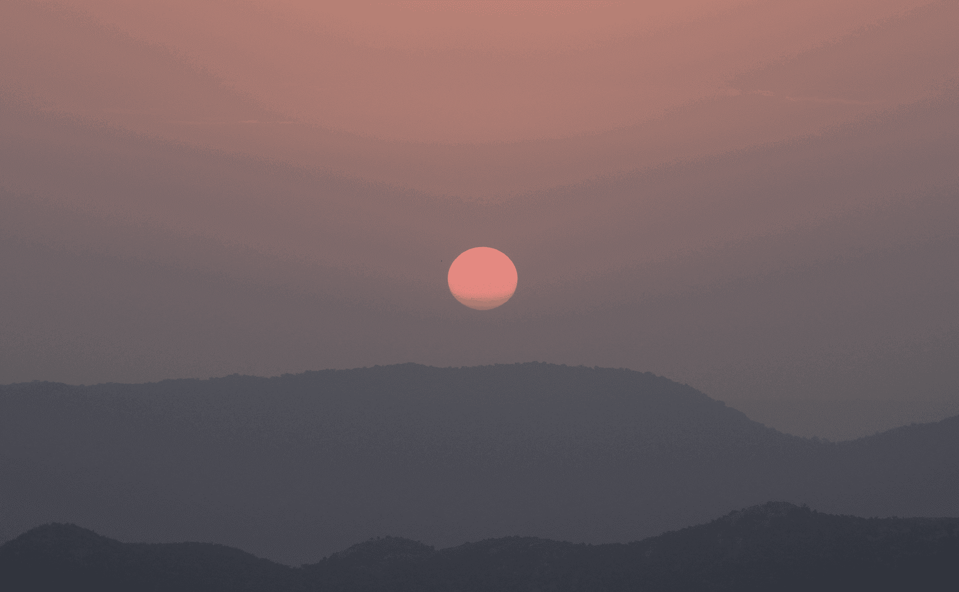 Sanft rosafarbene Sonne geht durch einen dunstigen Himmel über gestaffelten, silhouettenhaften Bergketten unter