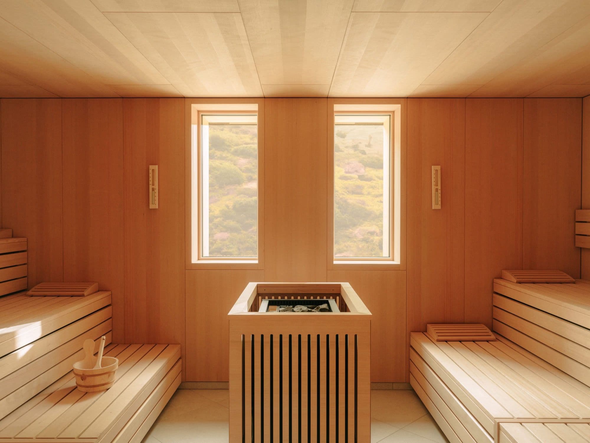 Holzverkleidetes Sauna-Interieur mit zwei hohen Fenstern zu grünen Hügeln und einem kleinen Holzkübel auf der Bank