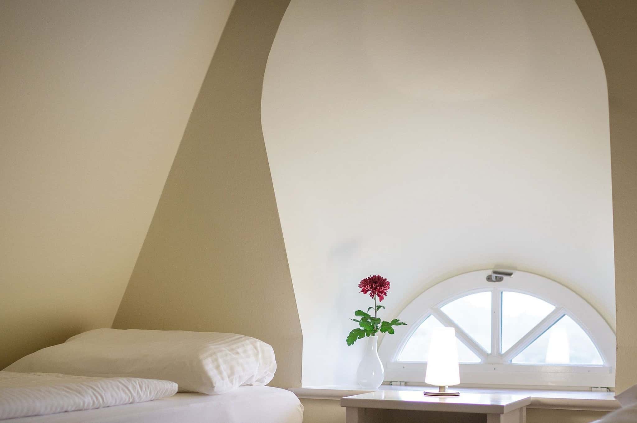Schlafzimmer unter der Dachschräge mit Rundbogenfenster, Nachttisch mit Lampe und Blume