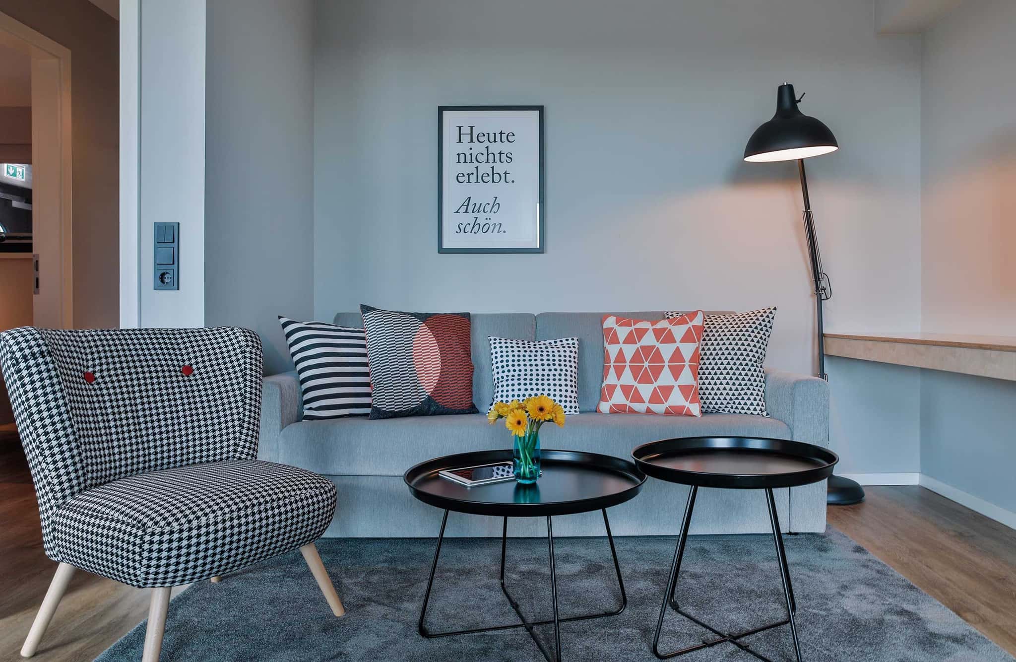 Wohnbereich mit grauem Sofa, Hahnentritt-Sessel, schwarzen Beistelltischen und Stehlampe, Bett & Bude