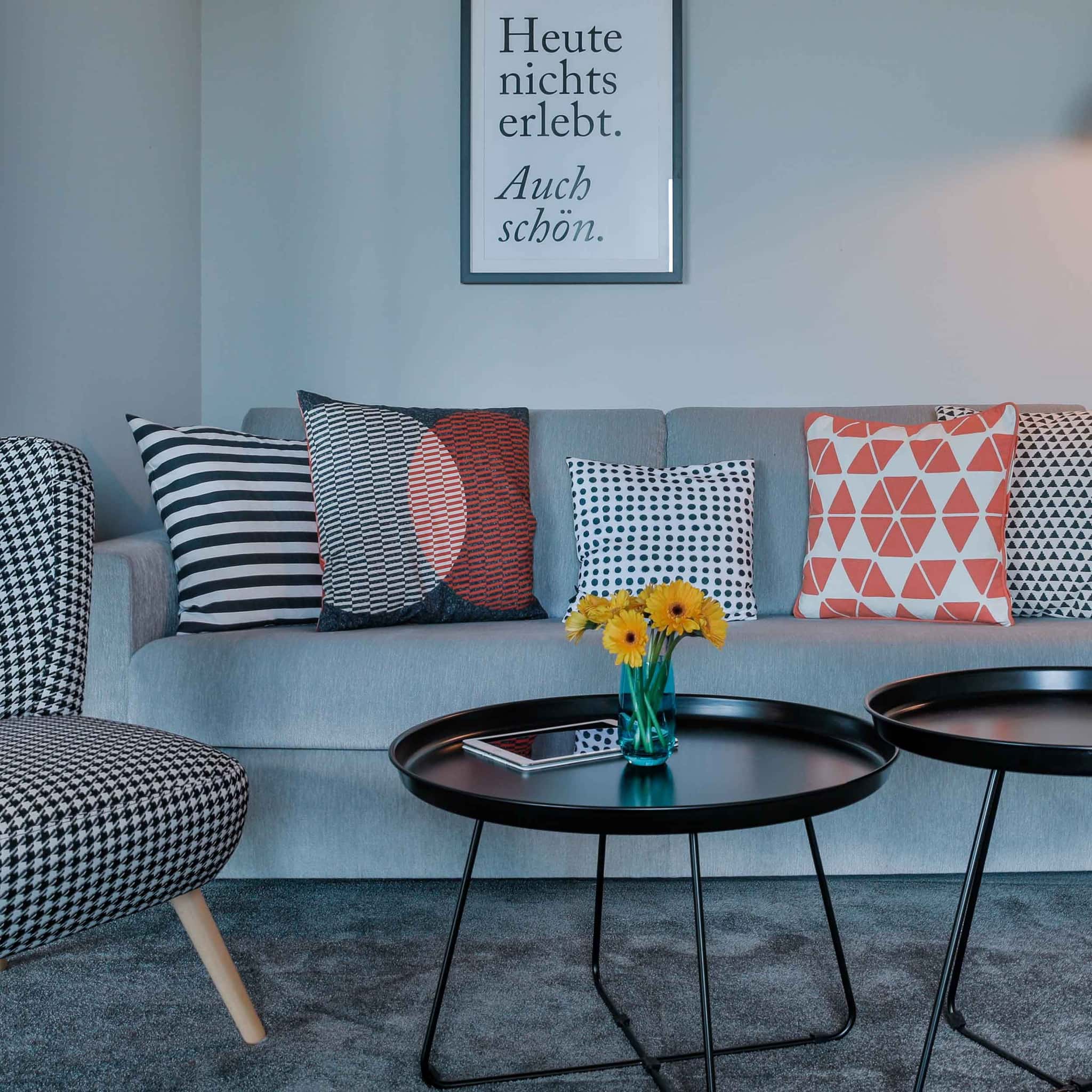 Graues Sofa mit gemusterten Kissen, Spruchbild, Hahnentritt-Sessel und schwarzen Beistelltischen, Bett & Bude