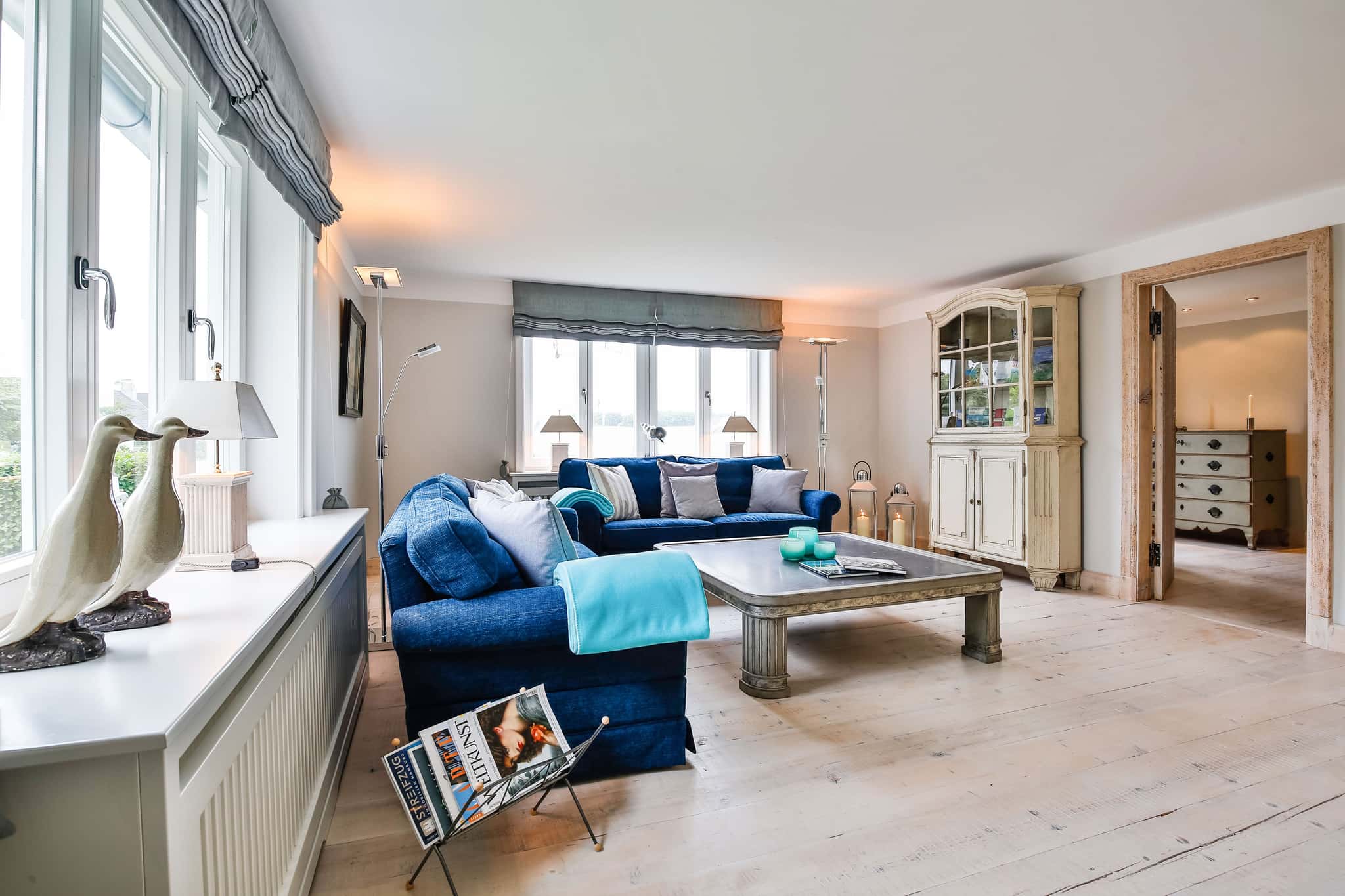 Wohnzimmer mit blauen Polstermöbeln, Couchtisch, Vitrine und Holzdielenboden im Fährhüs