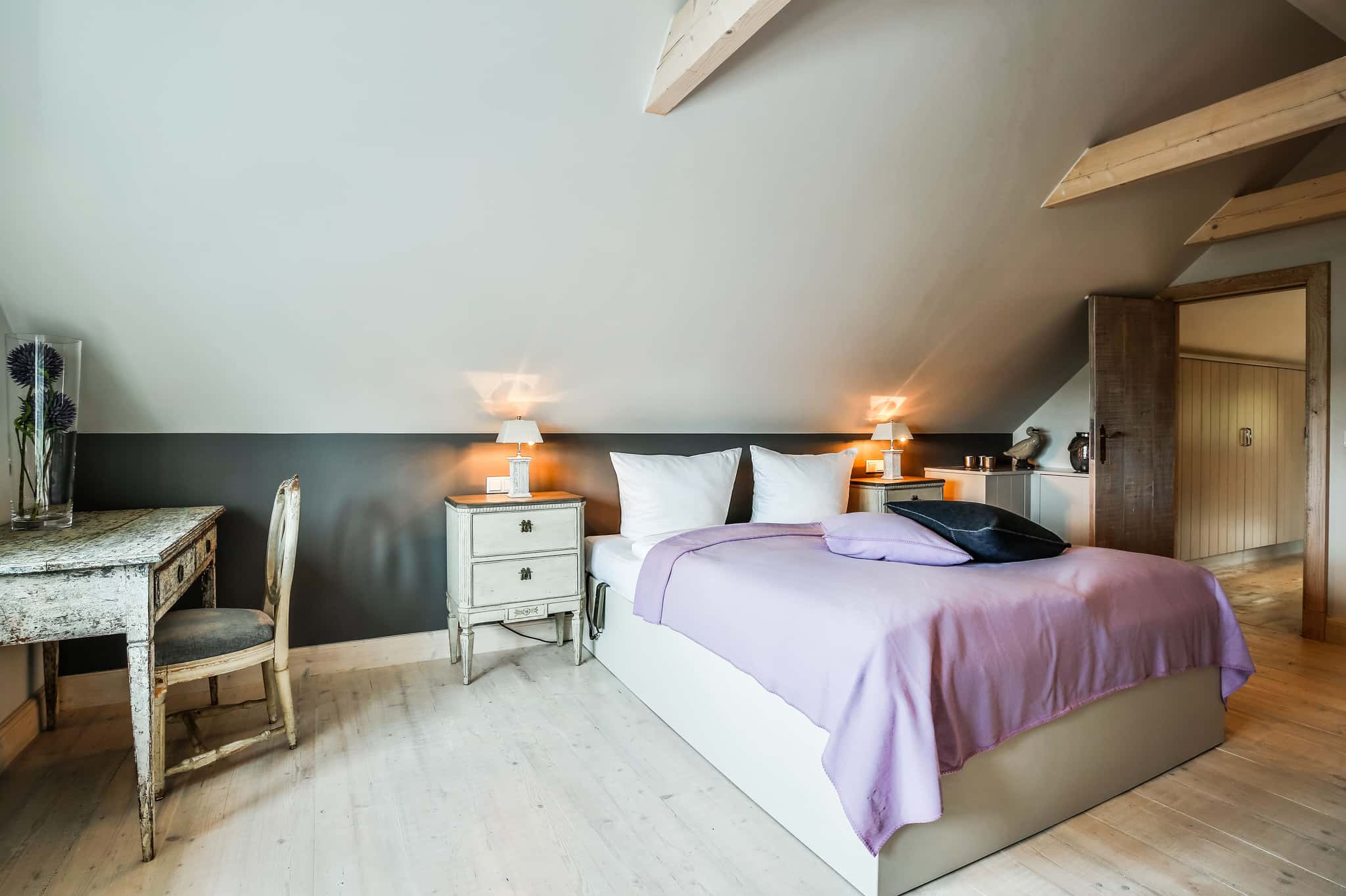 Schlafzimmer mit Bett unter der Dachschräge, fliederfarbener Tagesdecke, Nachttischen und Schreibtisch im Fährhüs