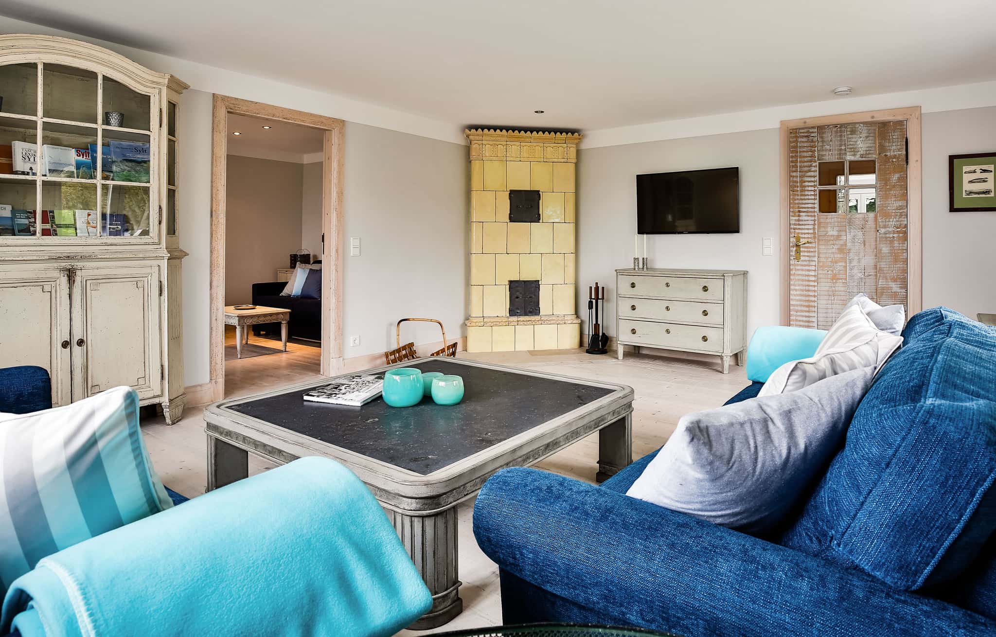 Wohnzimmer mit blauem Sofa, Couchtisch, gelbem Kachelofen und Vitrine im Fährhüs