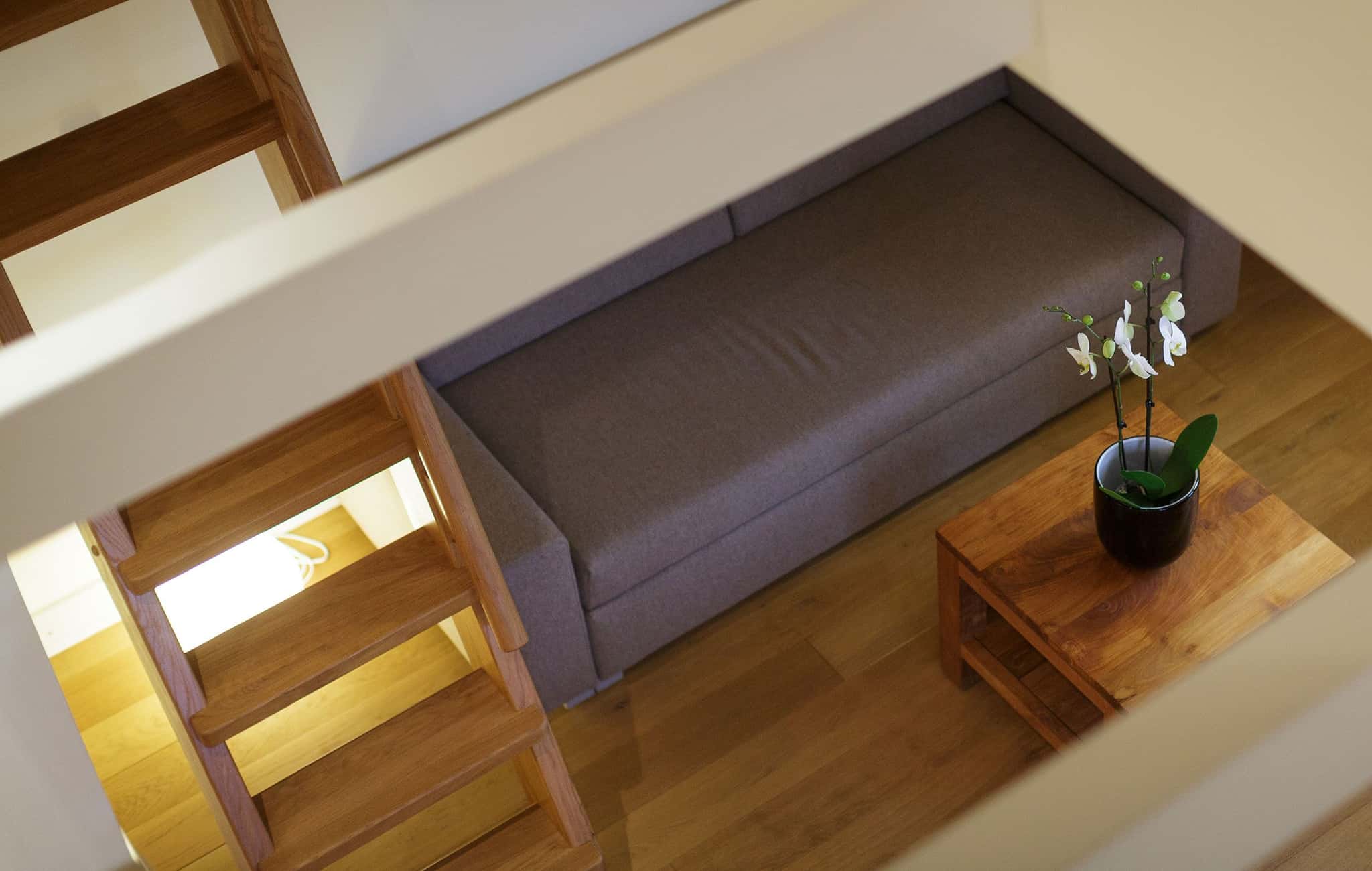 Blick von der Galerie auf ein Schlafsofa und einen Holztisch mit Orchidee, links die Holztreppe