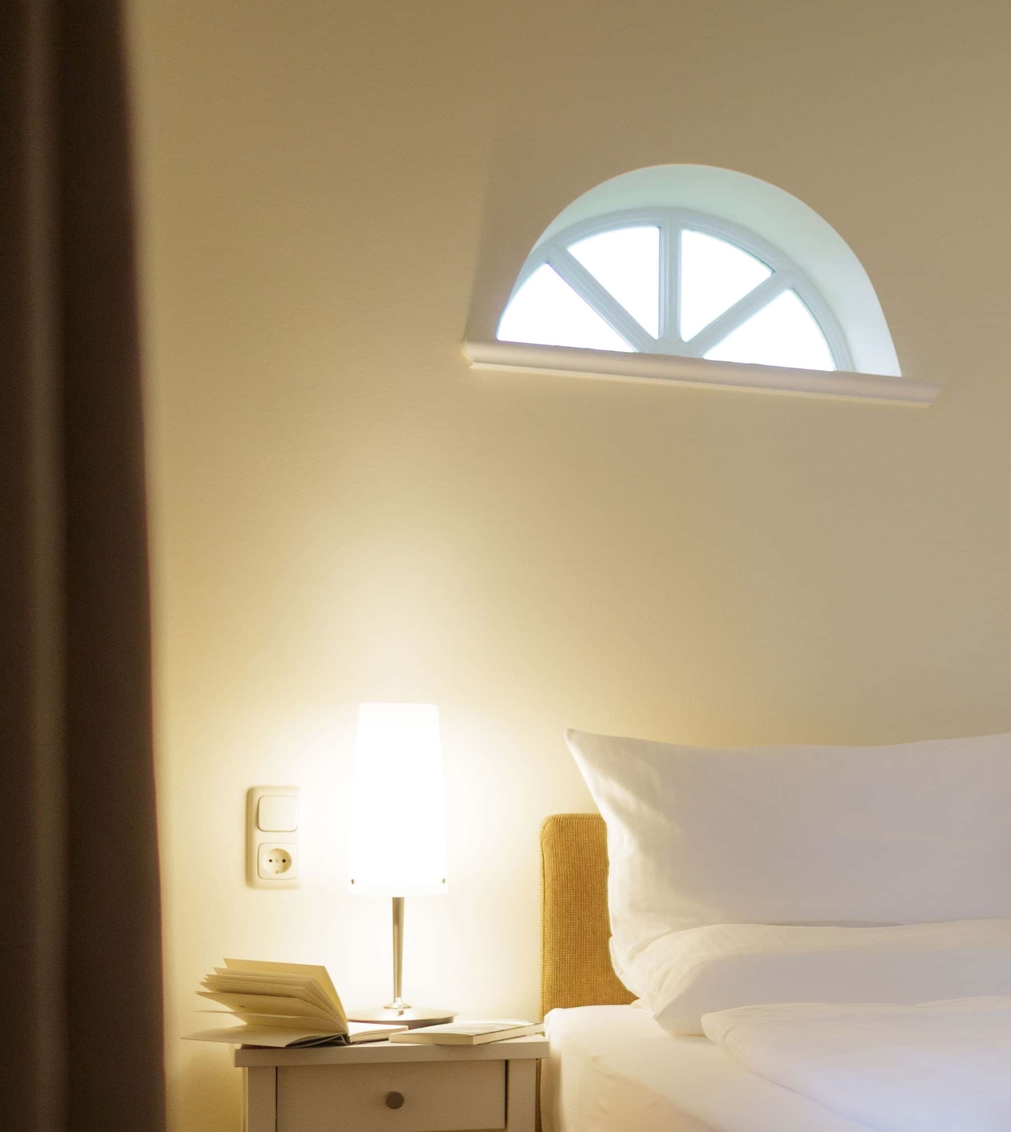 Nachttisch mit Leselampe und aufgeschlagenem Buch unter einem Halbrundfenster neben dem Bett