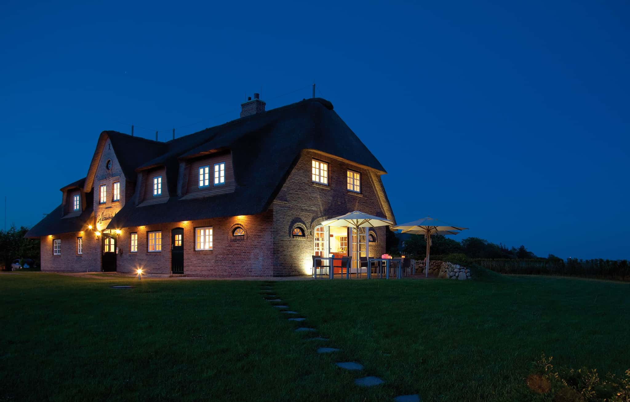 Reetgedecktes Backsteinhaus Lüüvhoog in der Dämmerung, beleuchtete Fenster, Terrasse und Rasen