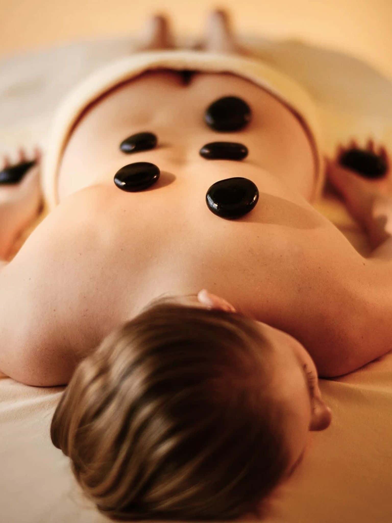 Eine Frau liegt bäuchlings auf der Massageliege, auf ihrem Rücken und in ihren Händen liegen dunkle, glatte Hot Stones.